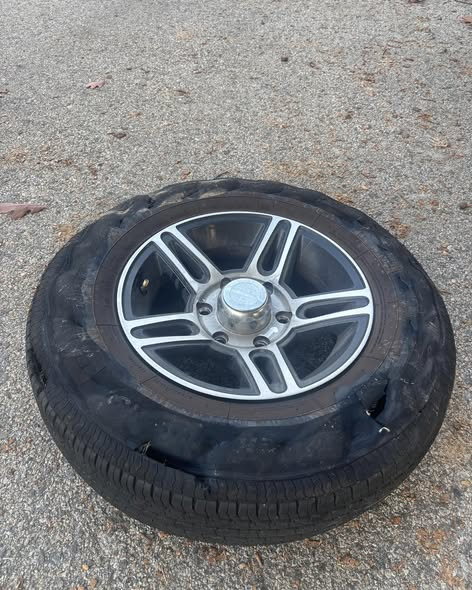 A shredded tire with its sidewall torn apart, still mounted on a five spoke machined alloy wheel, lying flat on an asphalt lot.