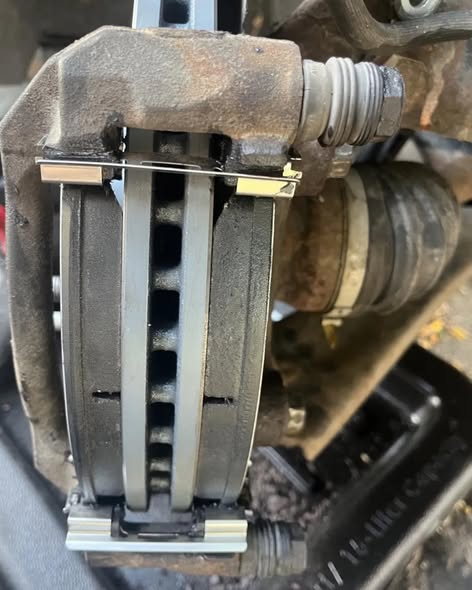 Close up of a rusty brake caliper off the rotor, with a pair of new black brake pads and bright new stainless hardware clips fitted into the bracket.