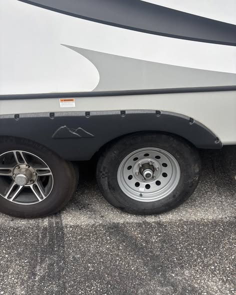 The side of a white RV on pavement showing both wheels of its tandem axle, an alloy wheel in front and a plain silver steel wheel at the rear.